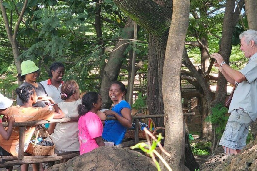 A group of people gathered outdoors, some sitting on a wooden platform, others standing, with trees and a stone structure in the background. 4