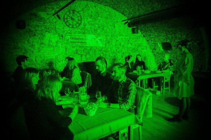 A group of people dining in a dimly lit, green-hued restaurant with a rustic brick wall and a clock. 1