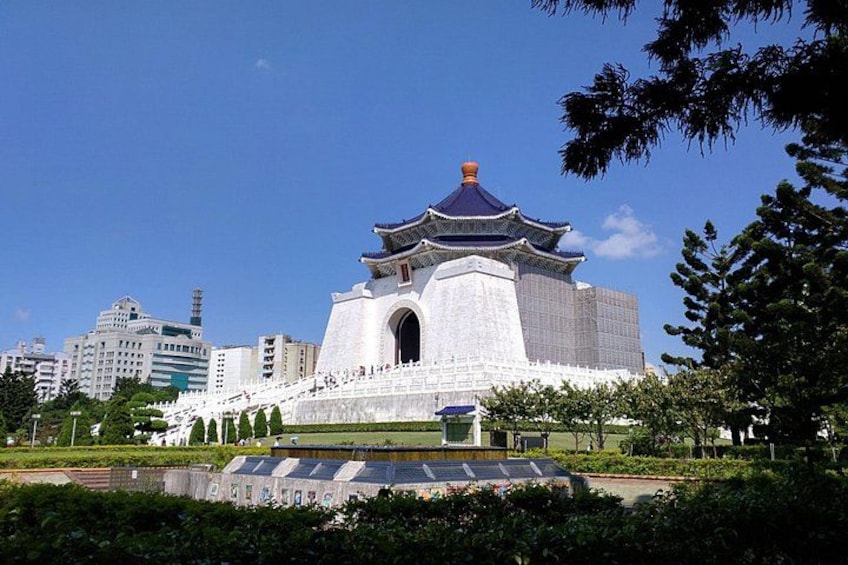 A traditional building with a blue roof and white walls, surrounded by greenery and modern buildings in the background. 4