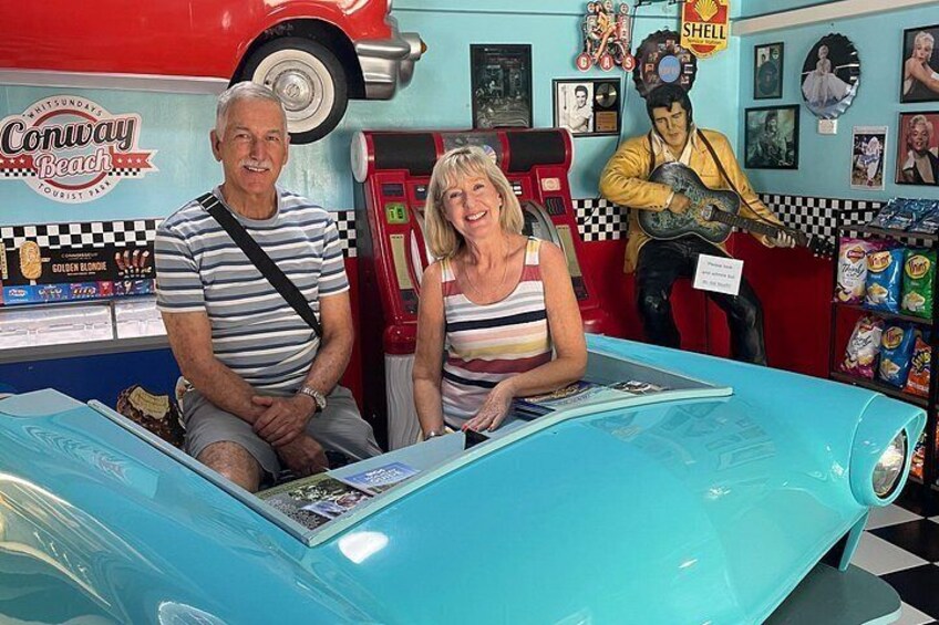 Exhibición de autos antiguos con un coche azul, un hombre y una mujer sentados en una mesa, y un músico tocando la guitarra. 1