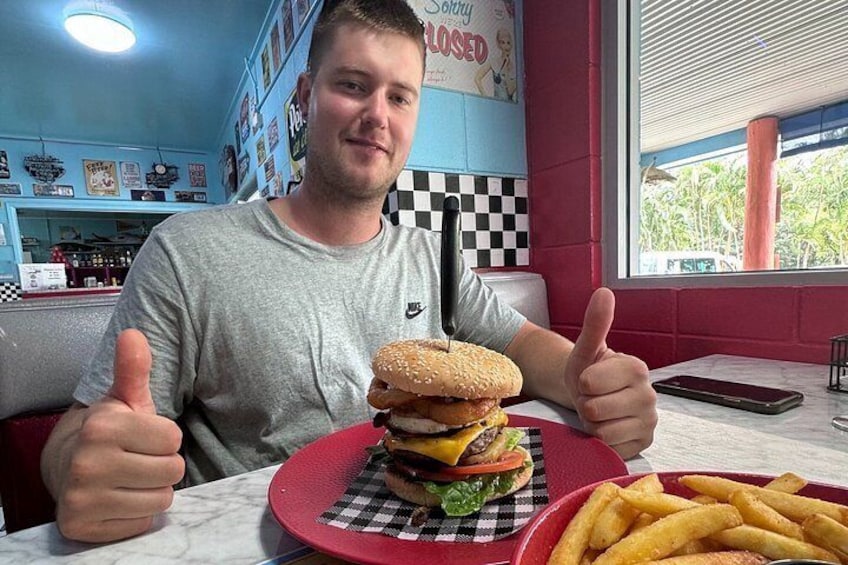 A person sitting at a table with a large burger and fries, a checkered tablecloth, and a 'Sorry, CLOSED' sign on the wall. 2
