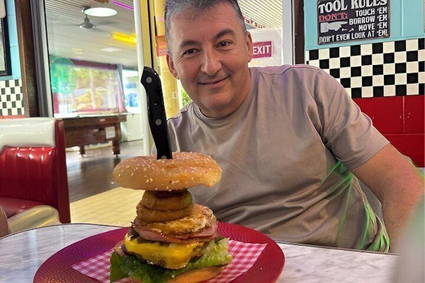 A person sitting at a table with a large burger and a knife on a checkered tablecloth. 3