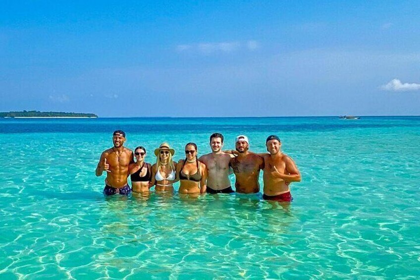 A group of people standing in shallow turquoise water with a clear sky and distant land visible. 1