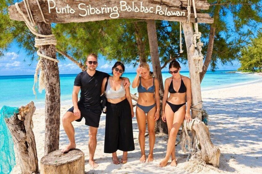 A wooden structure with the text 'Punta Sibaring Bolabac Polowan' on a beach with clear blue water and palm trees in the background. 2