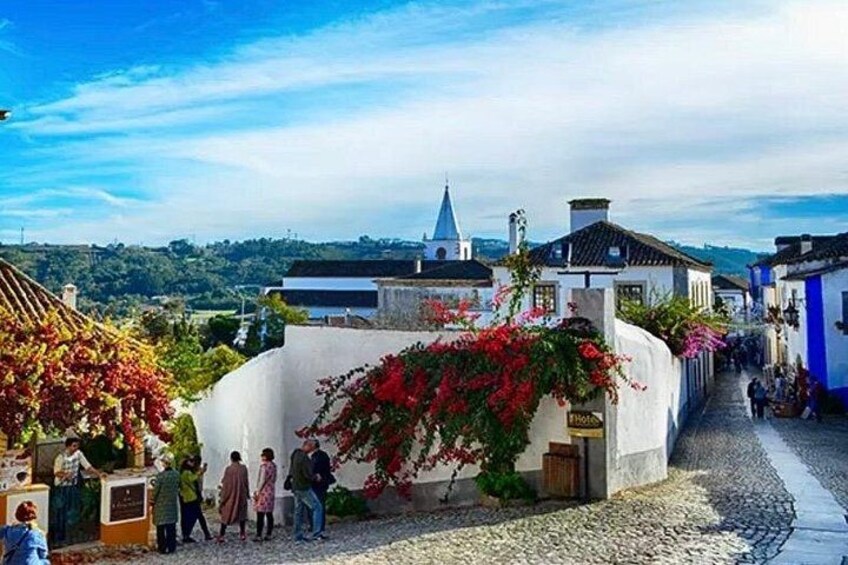 A cobblestone street with white buildings, red flowers, and a church spire in the background. 4