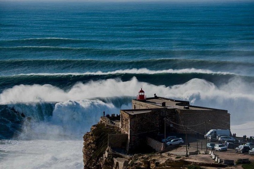 A coastal scene with waves crashing against a rocky outcrop, a lighthouse, and parked cars. 1