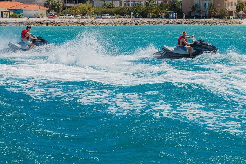 Two people jet skiing on a clear blue sea with buildings in the background. 3