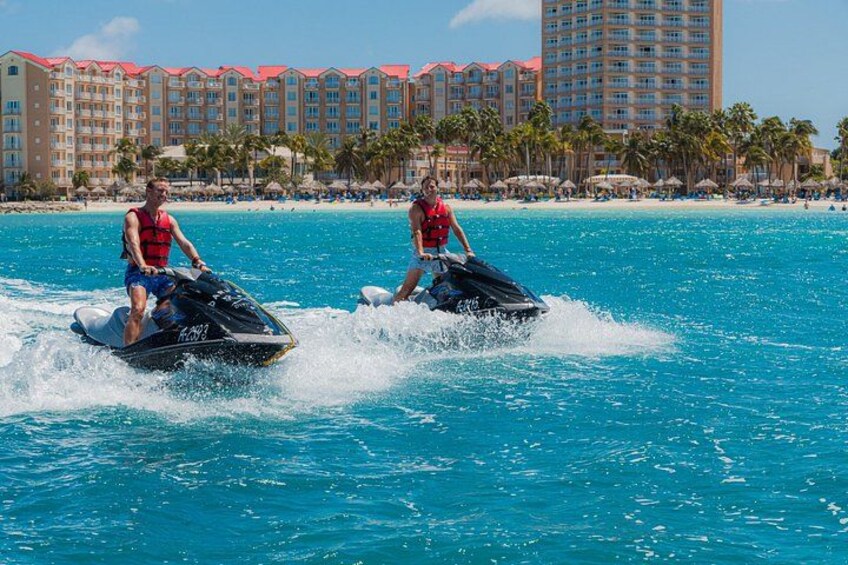 Two people jet skiing on a clear blue sea with a resort and palm trees in the background. 2