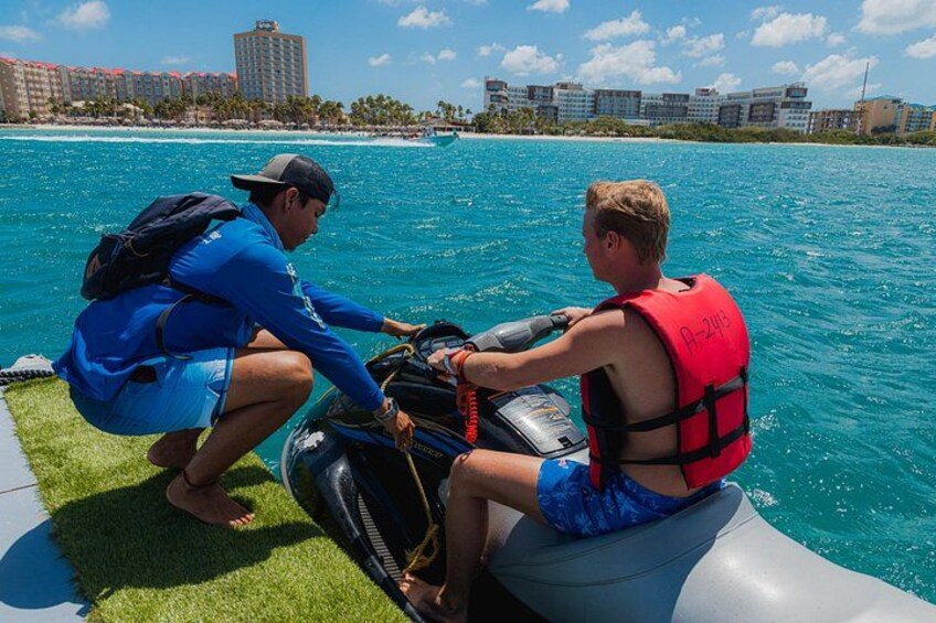 Two people on a jet ski, one wearing a red life jacket, with a cityscape and beach in the background. 4