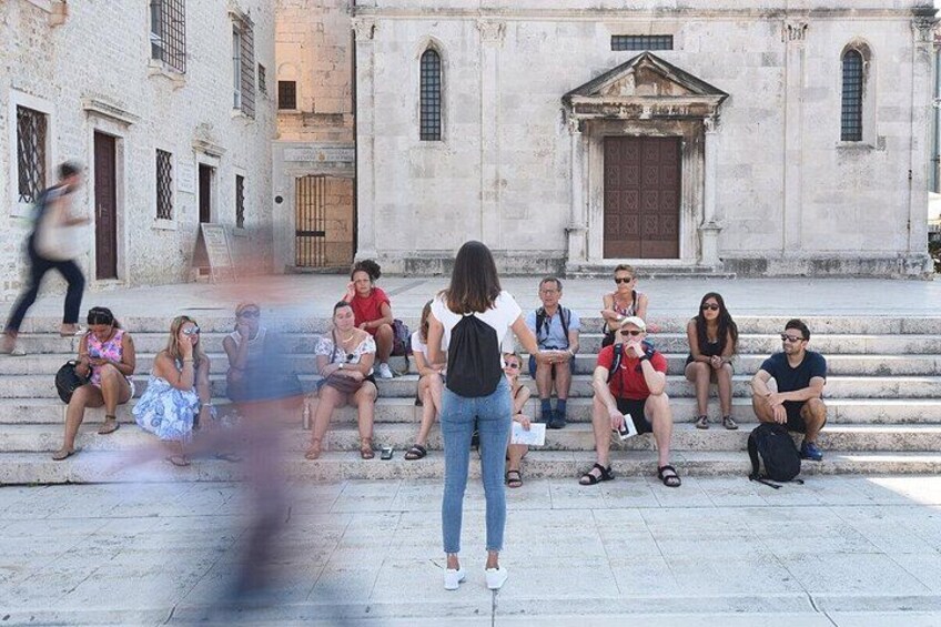 Un gruppo di persone sedute sugli scalini di fronte a un edificio storico con un'ampia porta in legno e finestre ad arco. 2