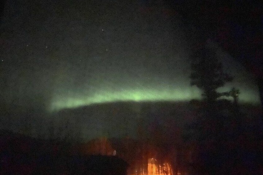 Escena nocturna con la aurora boreal, un bosque y un edificio distante. 2