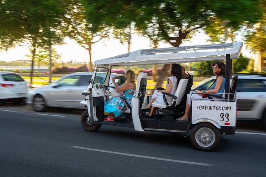 Un carrito de golf con tres pasajeros circulando por una calle, con coches estacionados y árboles al fondo. 2