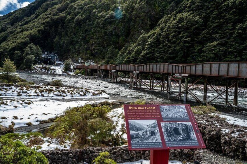 A snow-covered landscape with a wooden bridge and a sign about the Otira Rail Tunnel. 4