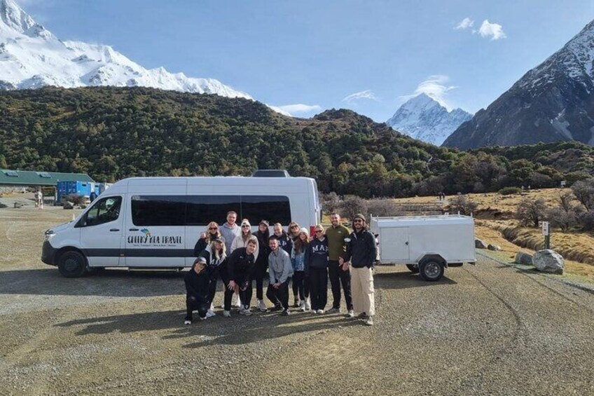 A group of people posing in front of a white van with a trailer, set against a mountainous landscape. 5