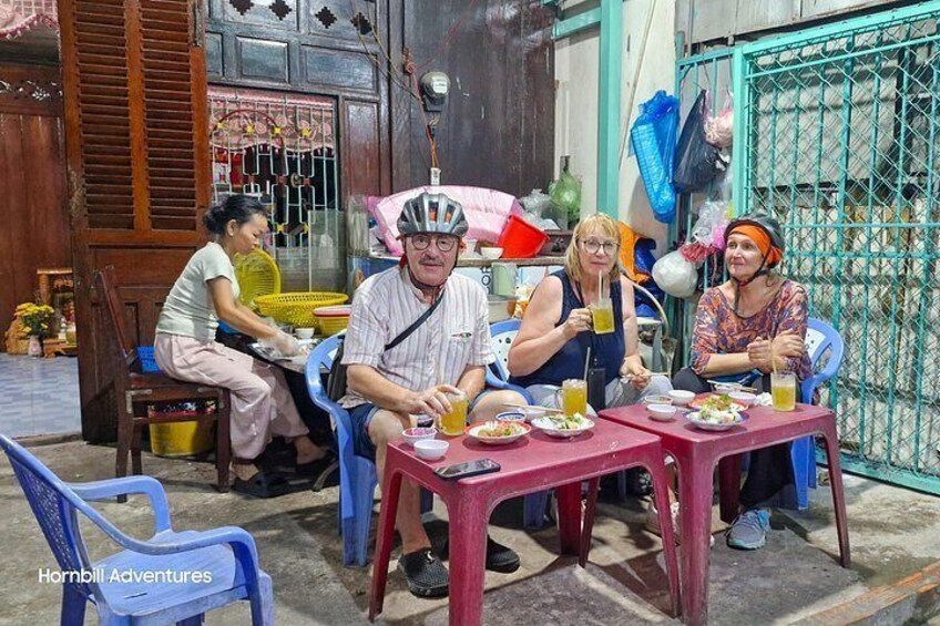 Four people sitting at a table with food and drinks, outdoors with a wooden door and a metal gate in the background. 3