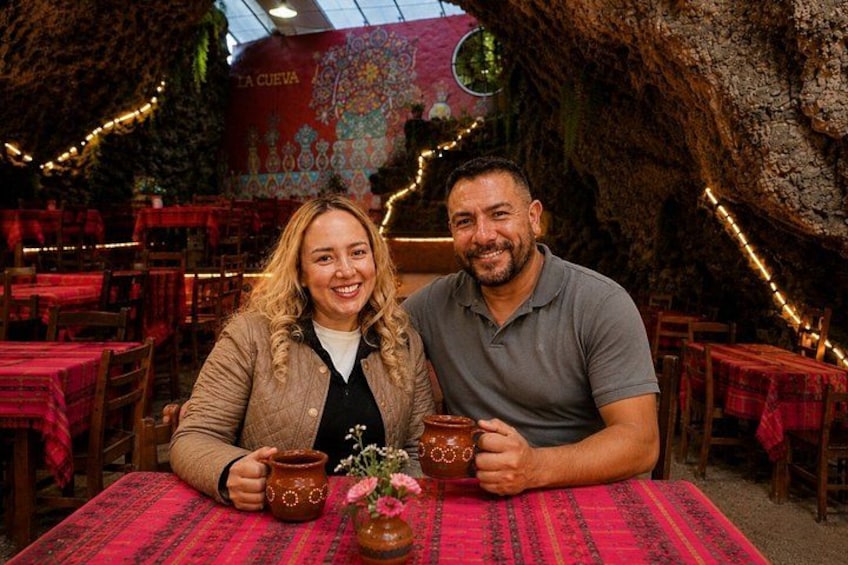 A couple seated at a table with a rustic interior, featuring a large tree trunk and a mural on the wall. 4