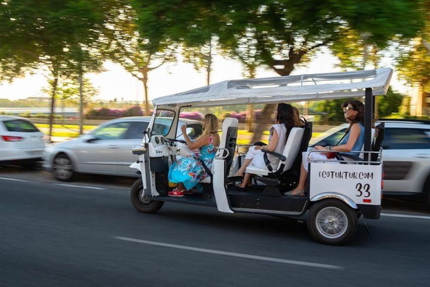 A golf cart with three passengers on a road with parked cars and trees in the background. 2