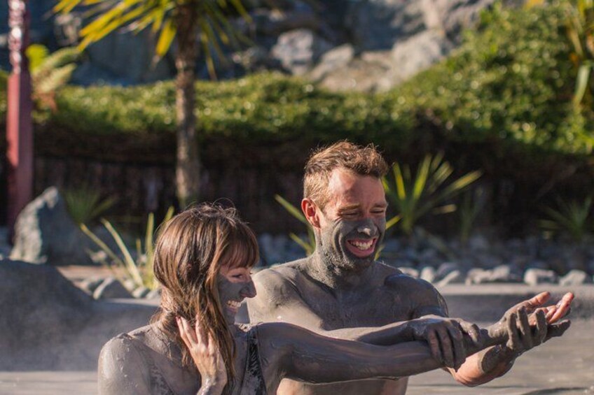 Two people covered in mud, standing outdoors with rocks and greenery in the background. 2