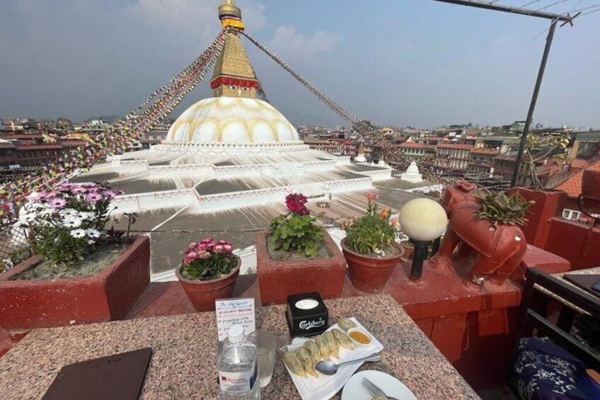 A rooftop terrace with potted plants, a table with food and drinks, and a view of a large white stupa with a golden spire. 3