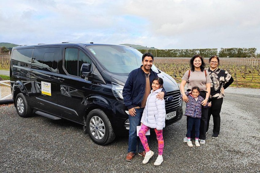 A family standing in front of a black van with a vineyard in the background. 3