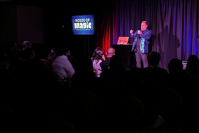 A magician performing on stage with a sign reading 'HOUSE OF MAGIC' in the background. 4