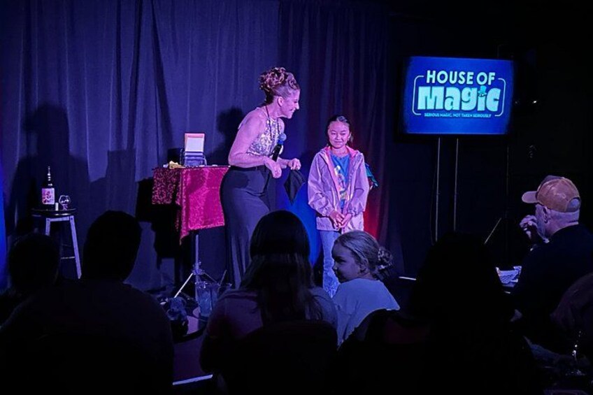 A stage with two people, a table with a laptop, and a sign that reads 'HOUSE OF MAGIC'. 3