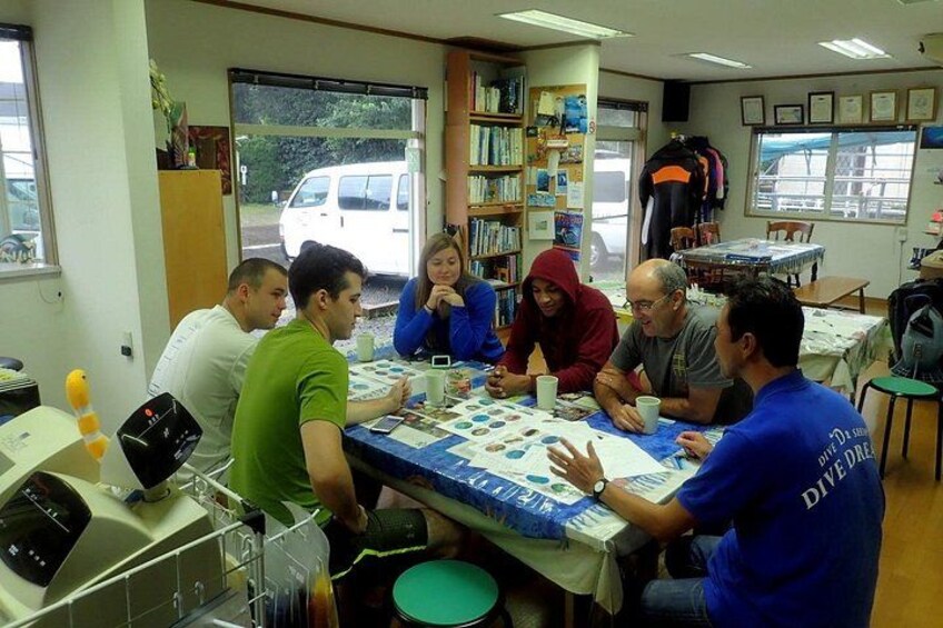 A group of people sitting around a table with a board game, in a room with bookshelves and a window. 2