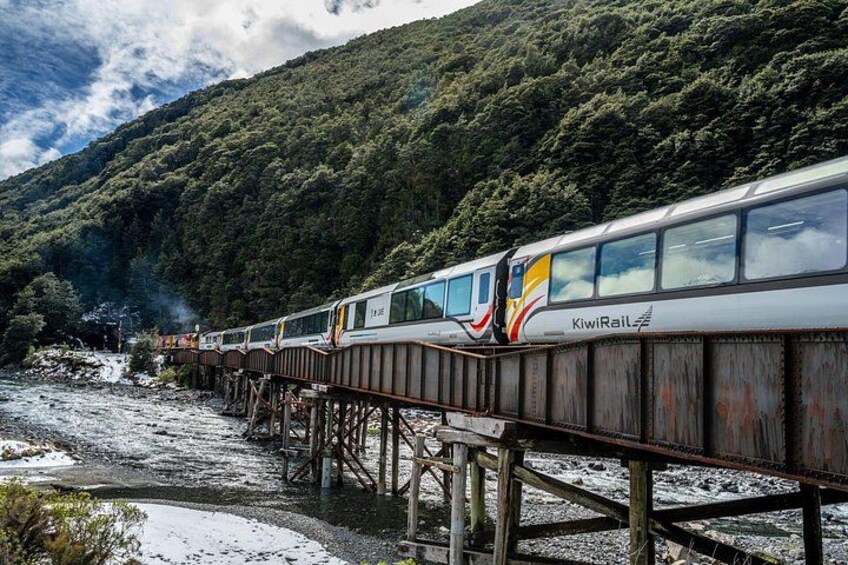 A train on a wooden bridge over a river, surrounded by forested hills. 2
