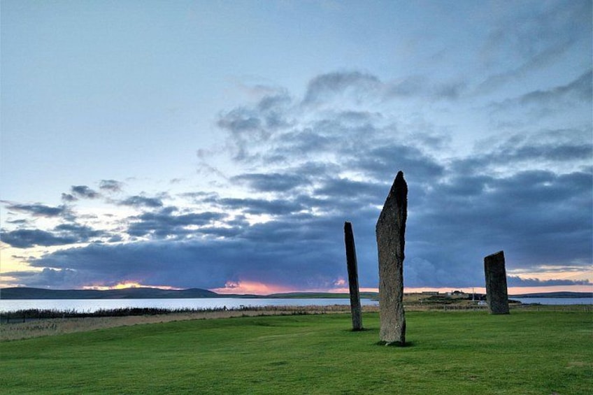 A grassy field with three standing stones and a sunset over the horizon. 4
