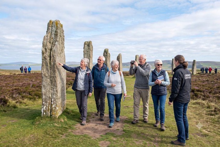 A group of people standing around ancient standing stones in a grassy field with a body of water and hills in the background. 5
