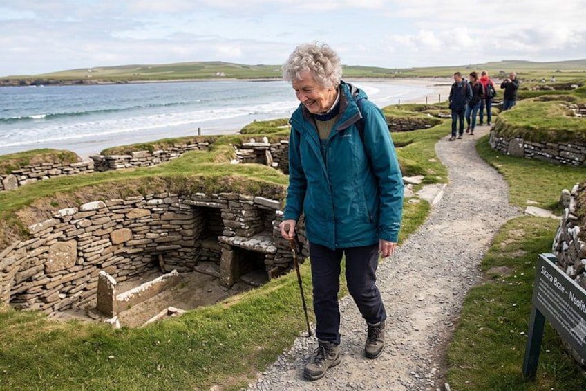 An elderly person walking on a gravel path near ancient stone structures by the sea. 3