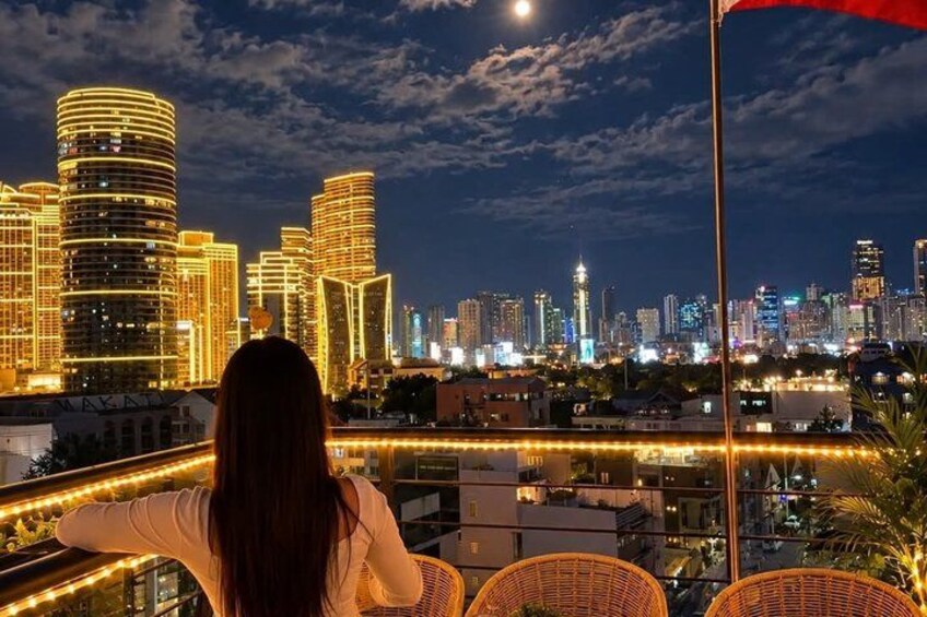 Un horizonte urbano nocturno con edificios iluminados y una persona sentada en un balcón. 1