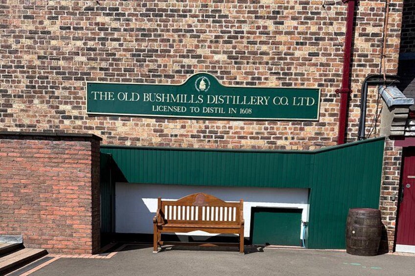 A brick building with a sign reading 'The Old Bushmills Distillery Co. Ltd' and a wooden bench in front. 3