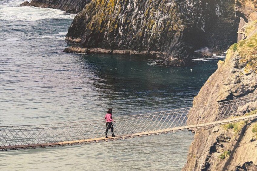 A person walking on a rope bridge over water, with rocky cliffs and ocean in the background. 2