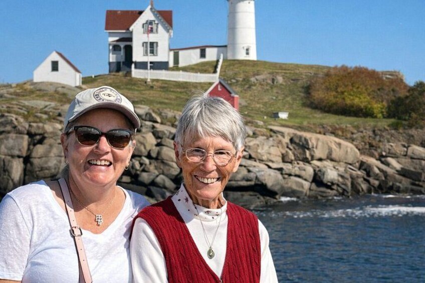 Two people standing in front of a lighthouse and a house on a rocky shore. 2