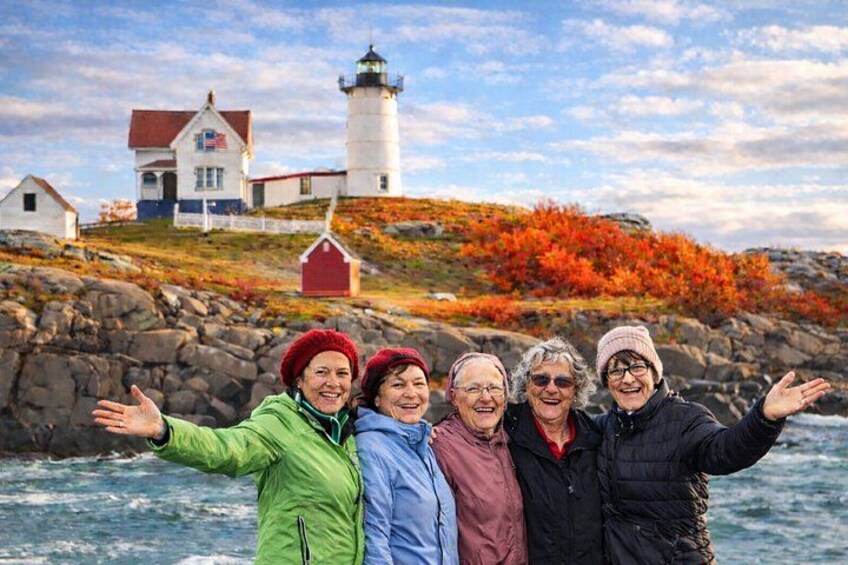 A group of people standing by a rocky shoreline with a lighthouse and buildings in the background. 1