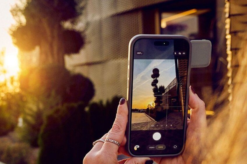 A person holding a smartphone with a camera app open, displaying a sunset scene with a building and trees. 2
