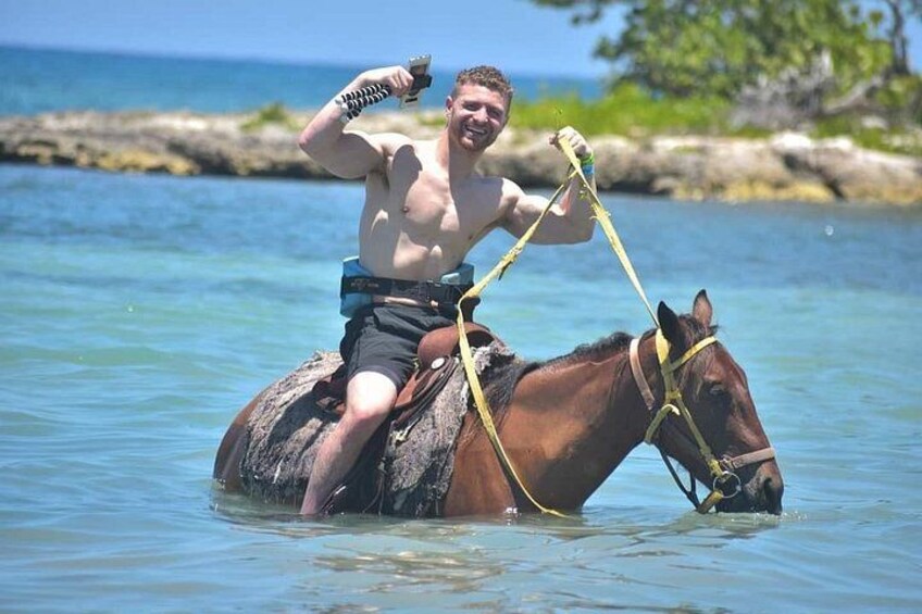A person riding a horse in shallow water with a rocky shoreline in the background. 3