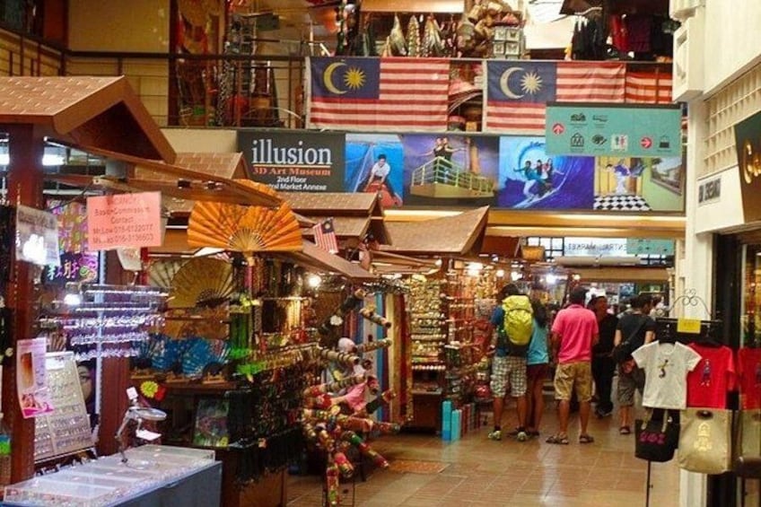 A bustling indoor market with various stalls selling goods, a 3D Art Museum sign, and Malaysian flags. 2