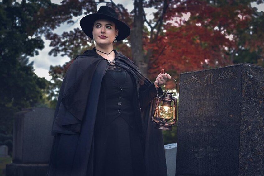 A person in a dark cloak and hat holding a lantern, standing next to a gravestone in a cemetery. 3