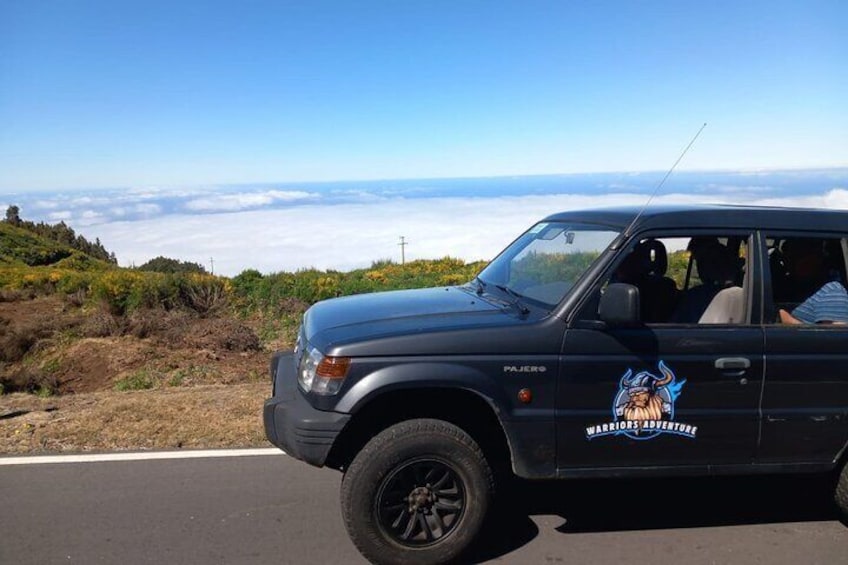 A black SUV with a 'Warriors Adventure' decal parked on a road with a scenic view of hills and clouds in the background. 5