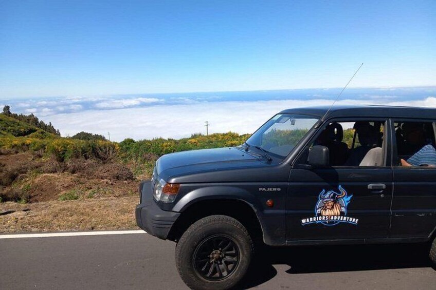 A black SUV with a 'Warriors Adventure' sticker parked on a road with a scenic view of mountains and clouds in the background. 4
