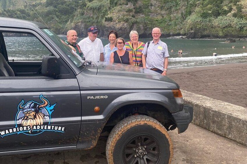 En gruppe mennesker står ved siden af en sort Pajero med et vikinge kriger logo, parkeret ved en strand hvor folk bader i vandet. 5