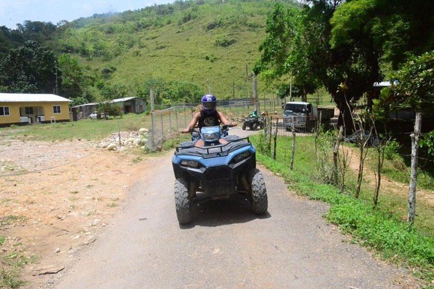 A person riding an ATV on a rural road with houses and greenery in the background. 2