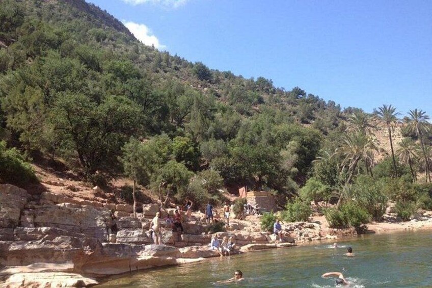 A rocky beach with people swimming and walking, surrounded by dense green trees and hills. 4