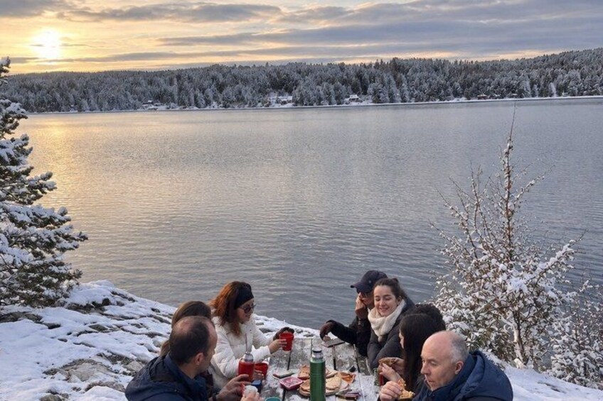 A group of people dining outdoors by a lake with snow-covered trees and a sunset in the background. 4