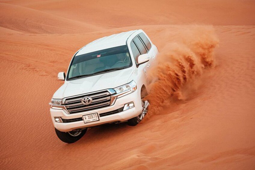 A white SUV driving through a desert with sand being kicked up by its tires. 1