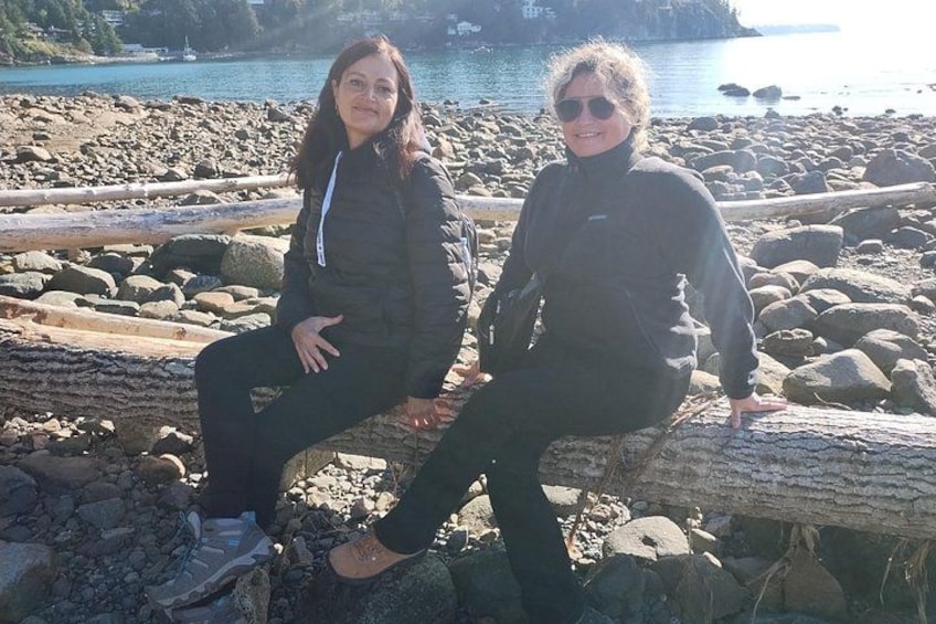 Due persone sedute su un tronco di legno lungo una riva rocciosa, con un tratto d'acqua e un'isola in lontananza. 3