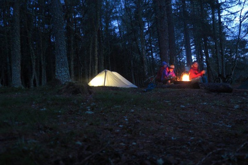 A tent in a forest at dusk, with a campfire nearby. 2