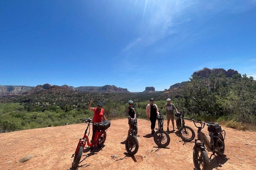 A group of cyclists on a dirt path with a scenic view of red rock formations and green vegetation under a clear sky. 1